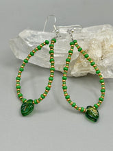 Load image into Gallery viewer, This pair of earrings features a dark green, pressed glass leaf charm in an iridescent finish that gives off subtle hues of gold, purple, blue, and pink. Complemented with green and gold glass beaded hoop earrings, this set is sure to sparkle.