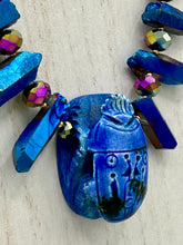 Load image into Gallery viewer, Powerful statement piece necklace features an eye-catching hand sculpted dark blue clay scarab beetle pendant. Adorned with vibrant sapphire luster quartz nuggets and sapphire faceted glass beaded necklace, this dramatic necklace artfully wraps around the neck, conveying an elegant and bold style. The Egyptian scarab symbol of rebirth, regeneration, and protection in the afterlife. Secure toggle clasp - Necklace is 23" length