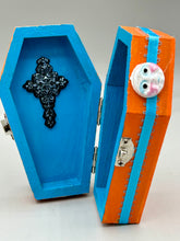 Load image into Gallery viewer, Coffin keepsake box represents the eternal love for a special kitty cat in your life. Cat silhouette sitting admiring the moon and stars. Day Of The Dead art was created for the cat lovers who adore their sweet kitty. Coffin-shaped box features a bright and vibrant orange exterior, with an adoring blue crescent moon and yellow star. Complemented with a shimmery blue ribbon and smiling moon, the inside is painted in a bright blue hue with black floral cross and detailing a traditional Mexican-themed fabric.
