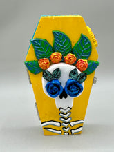 Load image into Gallery viewer, This coffin-shaped box features a bright canary yellow exterior, showcasing a vibrant Frida Kahlo inspired calavera. This beautifully handcrafted calavera is adorned with Frida's iconic floral crown. Complemented with a shimmery pastel-blue ribbon, the inside is painted in a bright blue hue accompanied by a black and silver floral cross and a Day Of The Dead kitten. The interior is also finished with a traditional Mexican-themed fabric.