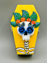 Load image into Gallery viewer, This coffin-shaped box features a bright canary yellow exterior, showcasing a vibrant Frida Kahlo inspired calavera. This beautifully handcrafted calavera is adorned with Frida's iconic floral crown. Complemented with a shimmery pastel-blue ribbon, the inside is painted in a bright blue hue accompanied by a black and silver floral cross and a Day Of The Dead kitten. The interior is also finished with a traditional Mexican-themed fabric.