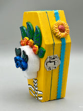 Load image into Gallery viewer, This coffin-shaped box features a bright canary yellow exterior, showcasing a vibrant Frida Kahlo inspired calavera. This beautifully handcrafted calavera is adorned with Frida's iconic floral crown. Complemented with a shimmery pastel-blue ribbon, the inside is painted in a bright blue hue accompanied by a black and silver floral cross and a Day Of The Dead kitten. The interior is also finished with a traditional Mexican-themed fabric.
