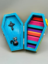 Load image into Gallery viewer, This coffin-shaped box features a bright canary yellow exterior, showcasing a vibrant Frida Kahlo inspired calavera. This beautifully handcrafted calavera is adorned with Frida's iconic floral crown. Complemented with a shimmery pastel-blue ribbon, the inside is painted in a bright blue hue accompanied by a black and silver floral cross and a Day Of The Dead kitten. The interior is also finished with a traditional Mexican-themed fabric.