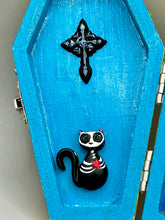 Load image into Gallery viewer, This coffin-shaped box features a bright canary yellow exterior, showcasing a vibrant Frida Kahlo inspired calavera. This beautifully handcrafted calavera is adorned with Frida's iconic floral crown. Complemented with a shimmery pastel-blue ribbon, the inside is painted in a bright blue hue accompanied by a black and silver floral cross and a Day Of The Dead kitten. The interior is also finished with a traditional Mexican-themed fabric.