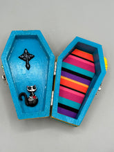 Load image into Gallery viewer, This coffin-shaped box features a bright canary yellow exterior, showcasing a vibrant Frida Kahlo inspired calavera. This beautifully handcrafted calavera is adorned with Frida's iconic floral crown. Complemented with a shimmery pastel-blue ribbon, the inside is painted in a bright blue hue accompanied by a black and silver floral cross and a Day Of The Dead kitten. The interior is also finished with a traditional Mexican-themed fabric.