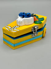 Load image into Gallery viewer, This coffin-shaped box features a bright canary yellow exterior, showcasing a vibrant Frida Kahlo inspired calavera. This beautifully handcrafted calavera is adorned with Frida's iconic floral crown. Complemented with a shimmery pastel-blue ribbon, the inside is painted in a bright blue hue accompanied by a black and silver floral cross and a Day Of The Dead kitten. The interior is also finished with a traditional Mexican-themed fabric.