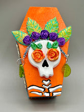 Load image into Gallery viewer, This coffin-shaped box features a bright and colorful exterior, showcasing a vibrant Frida Kahlo inspired calavera.  This beautifully handcrafted calavera is adorned with  Frida's iconic floral crown. To complete the look, the inside is also painted in colorful hues with a black and silver floral cross. The interior is also finished with a traditional Mexican-themed fabric.  L: 4 1/2“; W: 2 ½”; H: 1 5/8”