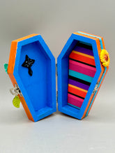 Load image into Gallery viewer, This coffin-shaped box features a bright and colorful exterior, showcasing a vibrant Frida Kahlo inspired calavera.  This beautifully handcrafted calavera is adorned with  Frida's iconic floral crown. To complete the look, the inside is also painted in colorful hues with a black and silver floral cross. The interior is also finished with a traditional Mexican-themed fabric.  L: 4 1/2“; W: 2 ½”; H: 1 5/8”
