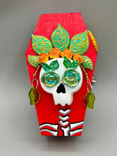 Load image into Gallery viewer, This coffin-shaped box features a bright and colorful exterior, showcasing a vibrant Frida Kahlo inspired calavera.  This beautifully handcrafted calavera is adorned with  Frida's iconic floral crown. To complete the look, the inside is also painted in colorful hues with a black and silver floral cross. The interior is also finished with a traditional Mexican-themed fabric.  L: 4 1/2“; W: 2 ½”; H: 1 5/8”