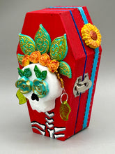Load image into Gallery viewer, This coffin-shaped box features a bright and colorful exterior, showcasing a vibrant Frida Kahlo inspired calavera.  This beautifully handcrafted calavera is adorned with  Frida's iconic floral crown. To complete the look, the inside is also painted in colorful hues with a black and silver floral cross. The interior is also finished with a traditional Mexican-themed fabric.  L: 4 1/2“; W: 2 ½”; H: 1 5/8”