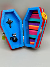 Load image into Gallery viewer, This coffin-shaped box features a bright and colorful exterior, showcasing a vibrant Frida Kahlo inspired calavera.  This beautifully handcrafted calavera is adorned with  Frida's iconic floral crown. To complete the look, the inside is also painted in colorful hues with a black and silver floral cross. The interior is also finished with a traditional Mexican-themed fabric.  L: 4 1/2“; W: 2 ½”; H: 1 5/8”