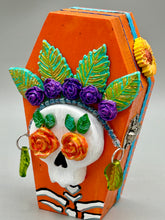 Load image into Gallery viewer, This coffin-shaped box features a bright and colorful exterior, showcasing a vibrant Frida Kahlo inspired calavera.  This beautifully handcrafted calavera is adorned with  Frida's iconic floral crown. To complete the look, the inside is also painted in colorful hues with a black and silver floral cross. The interior is also finished with a traditional Mexican-themed fabric.  L: 4 1/2“; W: 2 ½”; H: 1 5/8”