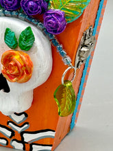 Load image into Gallery viewer, This coffin-shaped box features a bright and colorful exterior, showcasing a vibrant Frida Kahlo inspired calavera.  This beautifully handcrafted calavera is adorned with  Frida's iconic floral crown. To complete the look, the inside is also painted in colorful hues with a black and silver floral cross. The interior is also finished with a traditional Mexican-themed fabric.  L: 4 1/2“; W: 2 ½”; H: 1 5/8”