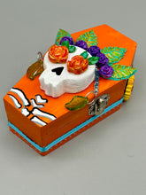 Load image into Gallery viewer, This coffin-shaped box features a bright and colorful exterior, showcasing a vibrant Frida Kahlo inspired calavera.  This beautifully handcrafted calavera is adorned with  Frida's iconic floral crown. To complete the look, the inside is also painted in colorful hues with a black and silver floral cross. The interior is also finished with a traditional Mexican-themed fabric.  L: 4 1/2“; W: 2 ½”; H: 1 5/8”