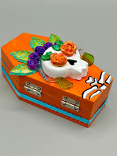 Load image into Gallery viewer, This coffin-shaped box features a bright and colorful exterior, showcasing a vibrant Frida Kahlo inspired calavera.  This beautifully handcrafted calavera is adorned with  Frida's iconic floral crown. To complete the look, the inside is also painted in colorful hues with a black and silver floral cross. The interior is also finished with a traditional Mexican-themed fabric.  L: 4 1/2“; W: 2 ½”; H: 1 5/8”