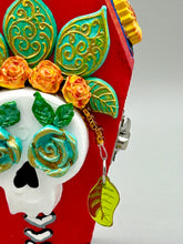 Load image into Gallery viewer, This coffin-shaped box features a bright and colorful exterior, showcasing a vibrant Frida Kahlo inspired calavera.  This beautifully handcrafted calavera is adorned with  Frida's iconic floral crown. To complete the look, the inside is also painted in colorful hues with a black and silver floral cross. The interior is also finished with a traditional Mexican-themed fabric.  L: 4 1/2“; W: 2 ½”; H: 1 5/8”