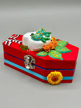 Load image into Gallery viewer, This coffin-shaped box features a bright and colorful exterior, showcasing a vibrant Frida Kahlo inspired calavera.  This beautifully handcrafted calavera is adorned with  Frida's iconic floral crown. To complete the look, the inside is also painted in colorful hues with a black and silver floral cross. The interior is also finished with a traditional Mexican-themed fabric.  L: 4 1/2“; W: 2 ½”; H: 1 5/8”