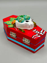 Load image into Gallery viewer, This coffin-shaped box features a bright and colorful exterior, showcasing a vibrant Frida Kahlo inspired calavera.  This beautifully handcrafted calavera is adorned with  Frida's iconic floral crown. To complete the look, the inside is also painted in colorful hues with a black and silver floral cross. The interior is also finished with a traditional Mexican-themed fabric.  L: 4 1/2“; W: 2 ½”; H: 1 5/8”