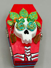 Load image into Gallery viewer, This coffin-shaped box features a bright and colorful exterior, showcasing a vibrant Frida Kahlo inspired calavera.  This beautifully handcrafted calavera is adorned with  Frida's iconic floral crown. To complete the look, the inside is also painted in colorful hues with a black and silver floral cross. The interior is also finished with a traditional Mexican-themed fabric.  L: 4 1/2“; W: 2 ½”; H: 1 5/8”