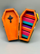 Load image into Gallery viewer, This coffin-shaped box features a bright and colorful exterior, showcasing a vibrant Frida Kahlo inspired calavera.  This beautifully handcrafted calavera is adorned with  Frida's iconic floral crown. To complete the look, the inside is also painted in colorful hues with a black and silver floral cross. The interior is also finished with a traditional Mexican-themed fabric.  L: 4 1/2“; W: 2 ½”; H: 1 5/8”