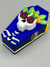 Load image into Gallery viewer, This coffin-shaped box features a bright and colorful exterior, showcasing a vibrant Frida Kahlo inspired calavera.  This beautifully handcrafted calavera is adorned with  Frida's iconic floral crown. To complete the look, the inside is also painted in colorful hues with a black and silver floral cross. The interior is also finished with a traditional Mexican-themed fabric.  L: 4 1/2“; W: 2 ½”; H: 1 5/8”