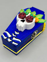 Load image into Gallery viewer, This coffin-shaped box features a bright and colorful exterior, showcasing a vibrant Frida Kahlo inspired calavera.  This beautifully handcrafted calavera is adorned with  Frida's iconic floral crown. To complete the look, the inside is also painted in colorful hues with a black and silver floral cross. The interior is also finished with a traditional Mexican-themed fabric.  L: 4 1/2“; W: 2 ½”; H: 1 5/8”