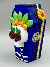 Load image into Gallery viewer, This coffin-shaped box features a bright and colorful exterior, showcasing a vibrant Frida Kahlo inspired calavera.  This beautifully handcrafted calavera is adorned with  Frida's iconic floral crown. To complete the look, the inside is also painted in colorful hues with a black and silver floral cross. The interior is also finished with a traditional Mexican-themed fabric.  L: 4 1/2“; W: 2 ½”; H: 1 5/8”