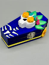 Load image into Gallery viewer, This coffin-shaped box features a bright and colorful exterior, showcasing a vibrant Frida Kahlo inspired calavera. This beautifully handcrafted calavera is adorned with Frida's iconic floral crown. To complete the look, the inside is also painted in colorful hues with a black and silver floral cross. The interior is also finished with a traditional Mexican-themed fabric. L: 4 1/2“; W: 2 ½”; H: 1 5/8”