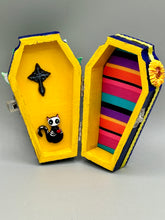 Load image into Gallery viewer, This coffin-shaped box features a bright and colorful exterior, showcasing a vibrant Frida Kahlo inspired calavera. This beautifully handcrafted calavera is adorned with Frida's iconic floral crown. To complete the look, the inside is also painted in colorful hues with a black and silver floral cross. The interior is also finished with a traditional Mexican-themed fabric. L: 4 1/2“; W: 2 ½”; H: 1 5/8”