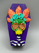 Load image into Gallery viewer, This coffin-shaped box features a bright and colorful exterior, showcasing a vibrant Frida Kahlo inspired calavera.  This beautifully handcrafted calavera is adorned with  Frida's iconic floral crown. To complete the look, the inside is also painted in colorful hues with a black and silver floral cross. The interior is also finished with a traditional Mexican-themed fabric.  L: 4 1/2“; W: 2 ½”; H: 1 5/8”