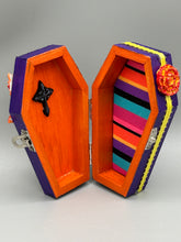 Load image into Gallery viewer, This coffin-shaped box features a bright and colorful exterior, showcasing a vibrant Frida Kahlo inspired calavera.  This beautifully handcrafted calavera is adorned with  Frida's iconic floral crown. To complete the look, the inside is also painted in colorful hues with a black and silver floral cross. The interior is also finished with a traditional Mexican-themed fabric.  L: 4 1/2“; W: 2 ½”; H: 1 5/8”