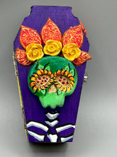 Load image into Gallery viewer, This coffin-shaped box features a bright and colorful exterior, showcasing a vibrant Frida Kahlo inspired calavera.  This beautifully handcrafted calavera is adorned with  Frida's iconic floral crown. To complete the look, the inside is also painted in colorful hues with a black and silver floral cross. The interior is also finished with a traditional Mexican-themed fabric.  L: 4 1/2“; W: 2 ½”; H: 1 5/8”