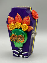 Load image into Gallery viewer, This coffin-shaped box features a bright and colorful exterior, showcasing a vibrant Frida Kahlo inspired calavera.  This beautifully handcrafted calavera is adorned with  Frida's iconic floral crown. To complete the look, the inside is also painted in colorful hues with a black and silver floral cross. The interior is also finished with a traditional Mexican-themed fabric.  L: 4 1/2“; W: 2 ½”; H: 1 5/8”
