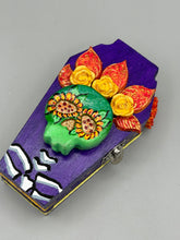 Load image into Gallery viewer, This coffin-shaped box features a bright and colorful exterior, showcasing a vibrant Frida Kahlo inspired calavera.  This beautifully handcrafted calavera is adorned with  Frida's iconic floral crown. To complete the look, the inside is also painted in colorful hues with a black and silver floral cross. The interior is also finished with a traditional Mexican-themed fabric.  L: 4 1/2“; W: 2 ½”; H: 1 5/8”