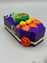 Load image into Gallery viewer, This coffin-shaped box features a bright and colorful exterior, showcasing a vibrant Frida Kahlo inspired calavera.  This beautifully handcrafted calavera is adorned with  Frida's iconic floral crown. To complete the look, the inside is also painted in colorful hues with a black and silver floral cross. The interior is also finished with a traditional Mexican-themed fabric.  L: 4 1/2“; W: 2 ½”; H: 1 5/8”
