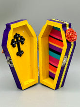 Load image into Gallery viewer, This coffin-shaped box features a bright and colorful exterior, showcasing a vibrant Frida Kahlo inspired calavera.  This beautifully handcrafted calavera is adorned with  Frida's iconic floral crown. To complete the look, the inside is also painted in colorful hues with a black and silver floral cross. The interior is also finished with a traditional Mexican-themed fabric.  L: 4 1/2“; W: 2 ½”; H: 1 5/8”