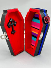 Load image into Gallery viewer, This coffin-shaped box features a bright and colorful exterior, showcasing a vibrant Frida Kahlo inspired calavera.  This beautifully handcrafted calavera is adorned with  Frida's iconic floral crown. To complete the look, the inside is also painted in colorful hues with a black and silver floral cross. The interior is also finished with a traditional Mexican-themed fabric.  L: 4 1/2“; W: 2 ½”; H: 1 5/8”