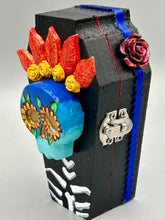 Load image into Gallery viewer, This coffin-shaped box features a bright and colorful exterior, showcasing a vibrant Frida Kahlo inspired calavera.  This beautifully handcrafted calavera is adorned with  Frida's iconic floral crown. To complete the look, the inside is also painted in colorful hues with a black and silver floral cross. The interior is also finished with a traditional Mexican-themed fabric.  L: 4 1/2“; W: 2 ½”; H: 1 5/8”