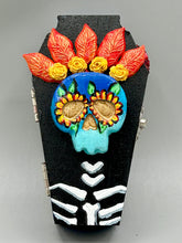 Load image into Gallery viewer, This coffin-shaped box features a bright and colorful exterior, showcasing a vibrant Frida Kahlo inspired calavera.  This beautifully handcrafted calavera is adorned with  Frida's iconic floral crown. To complete the look, the inside is also painted in colorful hues with a black and silver floral cross. The interior is also finished with a traditional Mexican-themed fabric.  L: 4 1/2“; W: 2 ½”; H: 1 5/8”