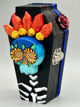 Load image into Gallery viewer, This coffin-shaped box features a bright and colorful exterior, showcasing a vibrant Frida Kahlo inspired calavera.  This beautifully handcrafted calavera is adorned with  Frida's iconic floral crown. To complete the look, the inside is also painted in colorful hues with a black and silver floral cross. The interior is also finished with a traditional Mexican-themed fabric.  L: 4 1/2“; W: 2 ½”; H: 1 5/8”