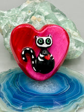 Load image into Gallery viewer, This hand-crafted art magnet is featuring a skeleton kitty cat, is the ideal home decor for cat lovers, Halloween, or Day Of The Dead enthusiasts. Perfect for adorning Day of the Dead ofrenda, a witchy altar, goth, or spooky Halloween home decor. Purrfectly cute and spooky Halloween magnet art to add to your fridge, locker, cubicle or toolbox. This hand-crafted heart-shaped magnet features a figurine of a black cat with ombre pink and red heart. 