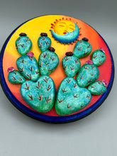 Load image into Gallery viewer, Southwest inspired wall art comes in a circle-shaped wood frame with a sun and crescent moon motifs. Its warm sky hues, including yellow, orange and dark red, are further complemented by hand sculpted cacti plants with cool green-blue tones with light and dark pink flowers. Diameter 6 3/4"