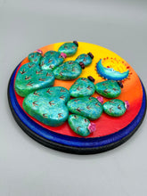 Load image into Gallery viewer, Southwest inspired wall art comes in a circle-shaped wood frame with a sun and crescent moon motifs. Its warm sky hues, including yellow, orange and dark red, are further complemented by hand sculpted cacti plants with cool green-blue tones with light and dark pink flowers. Diameter 6 3/4"