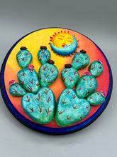 Load image into Gallery viewer, Southwest inspired wall art comes in a circle-shaped wood frame with a sun and crescent moon motifs. Its warm sky hues, including yellow, orange and dark red, are further complemented by hand sculpted cacti plants with cool green-blue tones with light and dark pink flowers. Diameter 6 3/4"