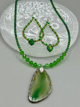 Load image into Gallery viewer, This pair of earrings features a dark green, pressed glass leaf charm in an iridescent finish that gives off subtle hues of gold, purple, blue, and pink. Complemented with green and gold glass beaded hoop earrings, this set is sure to sparkle.