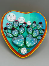 Load image into Gallery viewer, This handcrafted polymer clay wall art features a heart-shaped wood frame complimented with three smiling moon motifs.  Each moon symbolizing each of the realms of heavens, earth, and underworld. The complimentary color palette consists of cool sky hues, from a bright turquoise to a dark royal blue. The piece is further enhanced with hand sculpted cacti plants with blooming light and dark pink flowers.  The vibrant red and orange frame complements the cool-toned artwork.   H 7" x W 6 3/4” x D 1 1/4"
