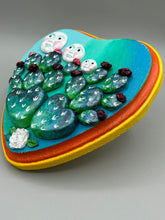 Load image into Gallery viewer, This handcrafted polymer clay wall art features a heart-shaped wood frame complimented with three smiling moon motifs.  Each moon symbolizing each of the realms of heavens, earth, and underworld. The complimentary color palette consists of cool sky hues, from a bright turquoise to a dark royal blue. The piece is further enhanced with hand sculpted cacti plants with blooming light and dark pink flowers.  The vibrant red and orange frame complements the cool-toned artwork.   H 7" x W 6 3/4” x D 1 1/4"