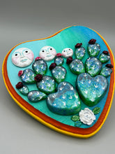 Load image into Gallery viewer, This handcrafted polymer clay wall art features a heart-shaped wood frame complimented with three smiling moon motifs.  Each moon symbolizing each of the realms of heavens, earth, and underworld. The complimentary color palette consists of cool sky hues, from a bright turquoise to a dark royal blue. The piece is further enhanced with hand sculpted cacti plants with blooming light and dark pink flowers.  The vibrant red and orange frame complements the cool-toned artwork.   H 7" x W 6 3/4” x D 1 1/4"