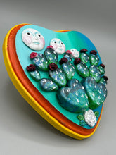 Load image into Gallery viewer, This handcrafted polymer clay wall art features a heart-shaped wood frame complimented with three smiling moon motifs.  Each moon symbolizing each of the realms of heavens, earth, and underworld. The complimentary color palette consists of cool sky hues, from a bright turquoise to a dark royal blue. The piece is further enhanced with hand sculpted cacti plants with blooming light and dark pink flowers.  The vibrant red and orange frame complements the cool-toned artwork.   H 7" x W 6 3/4” x D 1 1/4"