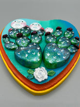 Load image into Gallery viewer, This handcrafted polymer clay wall art features a heart-shaped wood frame complimented with three smiling moon motifs.  Each moon symbolizing each of the realms of heavens, earth, and underworld. The complimentary color palette consists of cool sky hues, from a bright turquoise to a dark royal blue. The piece is further enhanced with hand sculpted cacti plants with blooming light and dark pink flowers.  The vibrant red and orange frame complements the cool-toned artwork.   H 7" x W 6 3/4” x D 1 1/4"